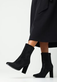 Botines negros con un upper texturizado y brillante, tacones cuadrados y cierre de cremallera lateral, llevados con una prenda negra.