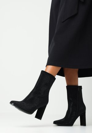 Bottines noires avec un matériau supérieur texturé et scintillant, des talons carrés et une fermeture éclair sur le côté, portées avec un vêtement noir.