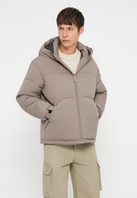 Jachetă puf din negru cu glugă, cu fermoar frontal, două buzunare laterale și un design matlasat. Purtați-o peste un pulover ușor și pantaloni bej.