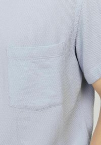 Lichtblauw gestructureerd shirt met korte mouwen, met een honingraatpatroon en een enkele borstzak. De stof lijkt zacht en ademend te zijn.
