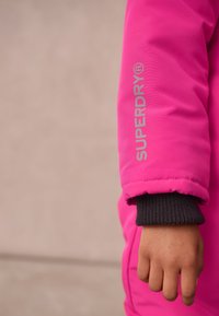 Rozā polsterētā virsjakas piedurkne ar pelēku "SUPERDRY" logo. Iezīmēts ar ribotu melnu aproci un gludu tekstūru. Redzama roka, kas tur audumu.