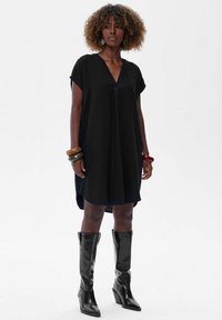 Czarna, luźna sukienka z dekoltem w serek, krótkimi rękawami i zaokrąglonym dół. Noszona z wysokimi, błyszczącymi czarnymi botkami oraz wieloma bransoletkami.