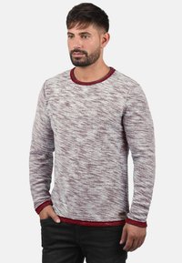Rød og hvit melert strikkegenser med rund hals og kontrastende rød trim ved mansjettene og nederste kant, kombinert med mørke jeans.