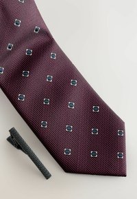 Burgunderfarbige Krawatte mit Muster, die eine Fischgrätenstruktur und quadratische blaue Akzente aufweist. Inklusive einer schwarzen Krawattennadel mit strukturierter Oberfläche.