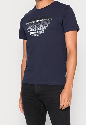 Mężczyzna noszący granatowy t-shirt z powtarzającym się napisem "JACK & JONES" i czarne dżinsy, stojący na jasnym, jednolitym tle.