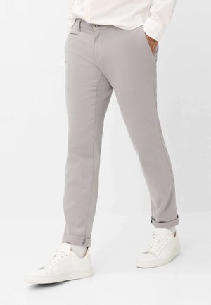 Mand iført lysegrå slim-fit bukser med op rullede manchetter, hvide sokker, hvide sneakers og en hvid langærmet skjorte med hænderne i lommerne.