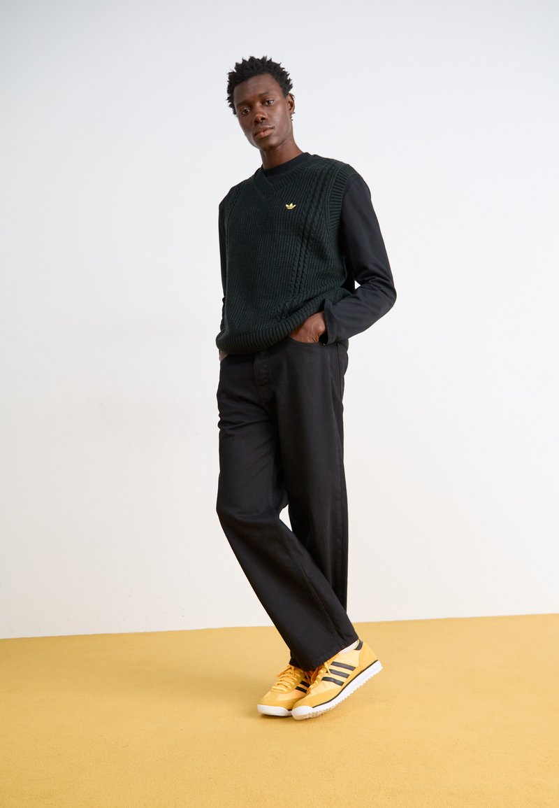 Schwarze weit geschnittene Hosen, schwarzes Langarmshirt unter einem grünen strukturierten Strickwesten mit einem goldenen Logo und gelben Sneakers mit schwarzen Streifen.