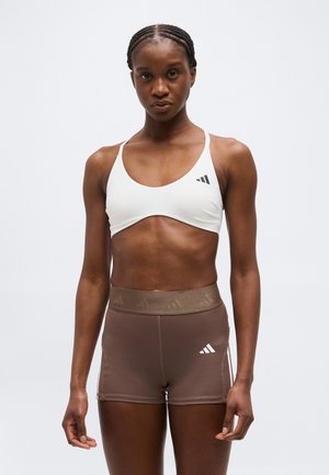 Atletyczna kobieta z zaplecionymi włosami, ubrana w biały sportowy biustonosz i brązowe, wysokowzrostne sportowe szorty, stojąca na jednolitym tle.