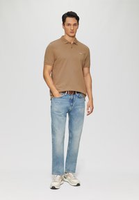 Camisa polo marrón, vaqueros azules, zapatillas claras. La camisa tiene un cuello clásico; los vaqueros son de corte relajado con una textura desgastada y corte recto.