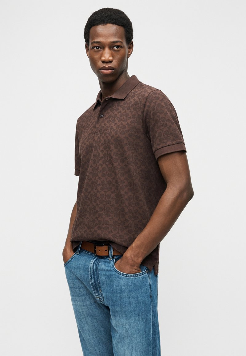 Young man standing with hands in pockets wearing a dark brown patterned polo shirt and blue jeans against a plain background.
