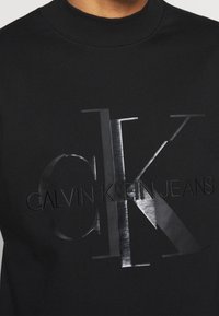 Felpa nera con grande logo CK lucido e testo "Calvin Klein Jeans" sul petto, indossata da una persona.