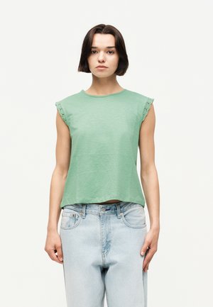 Jeune femme aux cheveux courts et foncés portant un top vert clair sans manches et un jean bleu clair, debout devant un fond uni.
