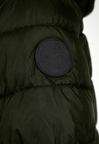 Doudoune vert foncé avec une texture brillante. Présente un logo noir circulaire avec les lettres "MNT" et "97" en relief. Les coutures sont visibles.