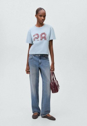 Junge Frau in hellblauem, tailliertem T-Shirt mit der Nummer 28, ausgewaschenen blauen Jeans, braunen, bespannten Ballerinas, die eineburgunderrote Handtasche hält.