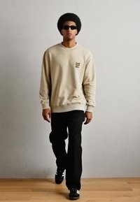 Felpa beige con un design di orso sul petto, abbinata a pantaloni neri e sneakers nere. Tessuto liscio, vestibilità rilassata.