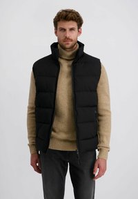 Schwarze Steppweste mit hohem Kragen, gestepptem Design und Reißverschluss, getragen über einem beigefarbenen Rollkragenpullover, kombiniert mit dunkelgrauen Hosen.
