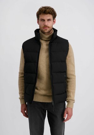 Schwarze Steppweste mit hohem Kragen, gestepptem Design und Reißverschluss, getragen über einem beigefarbenen Rollkragenpullover, kombiniert mit dunkelgrauen Hosen.