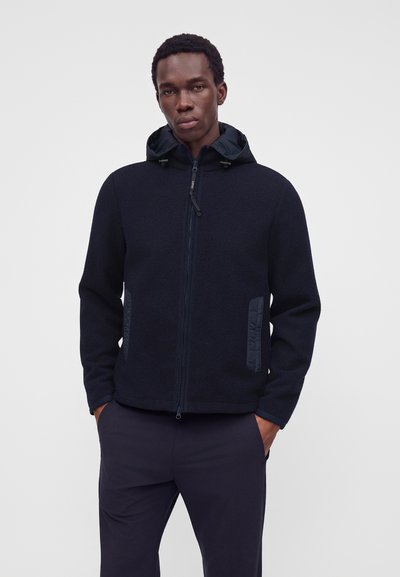 Homme portant une veste zippée bleu marine foncé avec capuche et les mains dans les poches sur un fond clair uni.