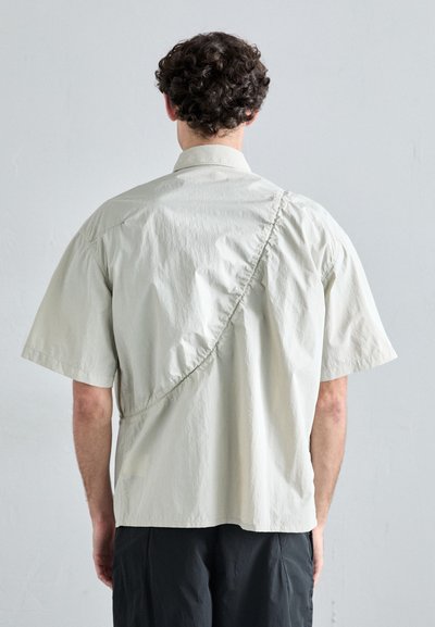 Chemise à manches courtes en beige clair, présentant un design drapé asymétrique avec des détails froncés au dos. Tissu doux et texturé.