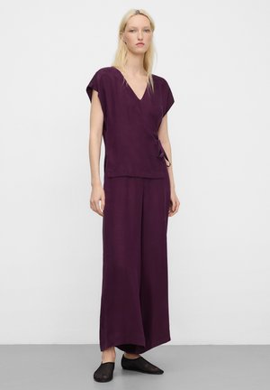 Donna con capelli biondi che indossa una maglia incrociata senza maniche di colore viola scuro e pantaloni larghi abbinati, con scarpe slip-on nere in rete.