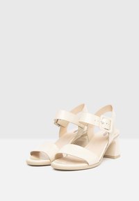 Sandales en cuir verni beige avec un talon épais, une bride de cheville réglable, un bout ouvert et une texture lisse.