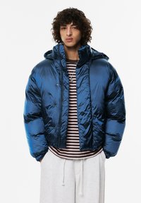 Kurtka puchowa w błyszczącym ciemnoniebieskim materiale, krótki krój, z kapturem i zapięciem na zatrzaski z przodu. Zestawiona z paskowaną koszulą pod spodem.