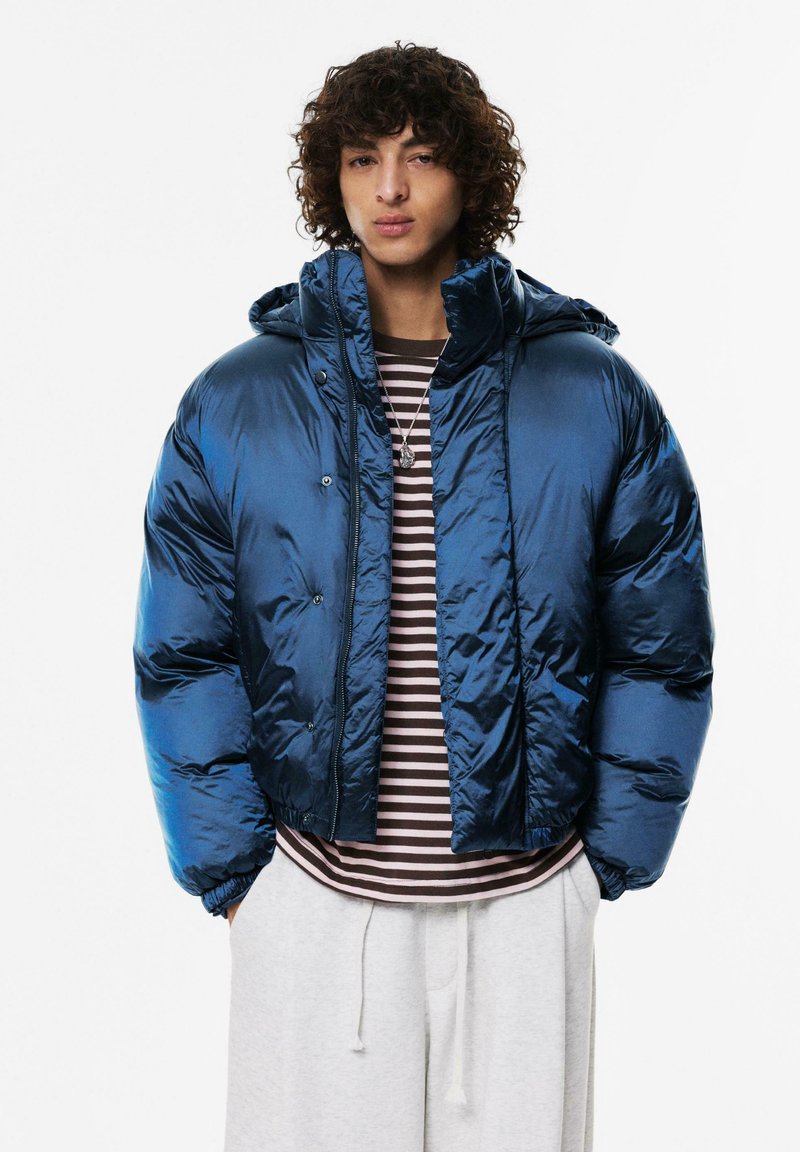 Opblaasjack van glanzend donkerblauw materiaal, cropped ontwerp, met een capuchon en voorste drukknoopsluiting. Gecombineerd met een gestreept shirt eronder.
