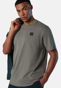 Graues T-Shirt mit kurzen Ärmeln und rundem Ausschnitt, mit einem schwarzen Logo-Patch auf der Brust. Es hat eine glatte Stofftextur und eine lockere Passform.