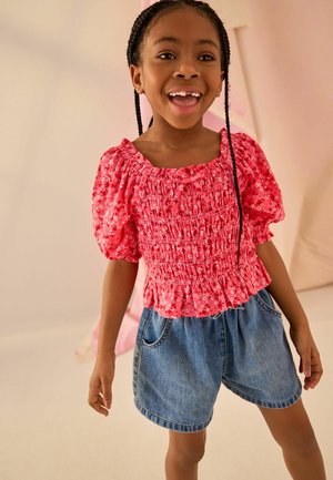 Lächelndes junges Mädchen mit geflochtenem Haar, das ein rosa florales Oberteil mit Puffärmeln und blaue Jeansshorts vor neutralem Hintergrund trägt.