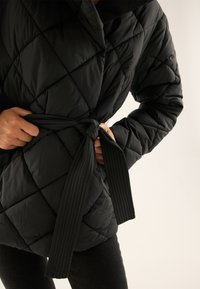 Osoba wiążąca pasek czarnej pikowanej kurtki, widoczna od klatki piersiowej do połowy uda, na jasnym neutralnym tle.