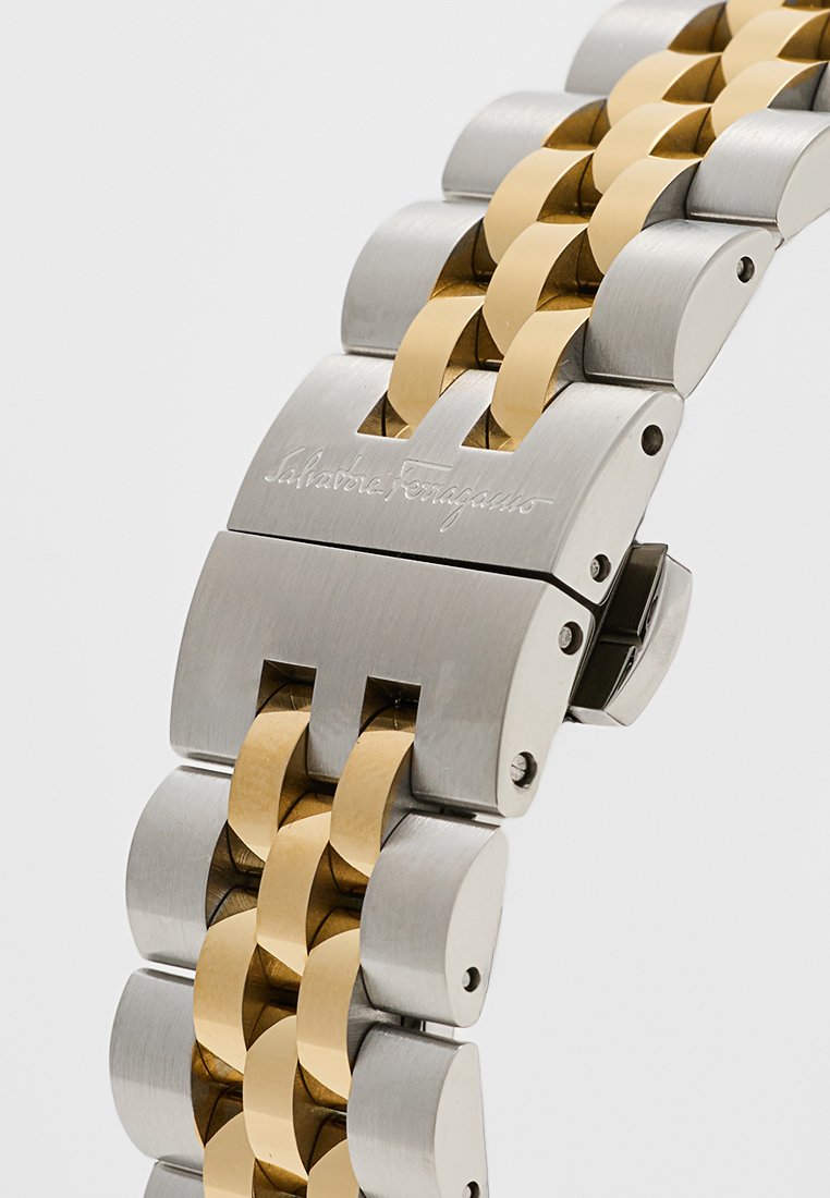 Bracelet en métal pour montre avec des maillons alternant entre acier brossé argenté et or poli. Comprend un fermoir avec détail de marque gravé.