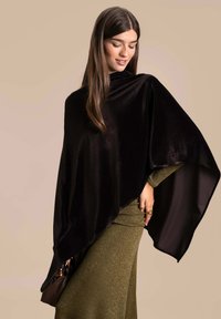 Poncho de terciopelo negro con un diseño de corte amplio y mangas drapeadas, llevado sobre un vestido brillante de color verde oliva. Textura suave y flujo elegante.