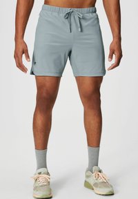 Helle graue Schwimmshorts mit einem elastischen Bund und Kordelzug, seitlichen Schlitzen und einem kleinen Logodetail. Kombiniert mit grauen Socken und Turnschuhen.