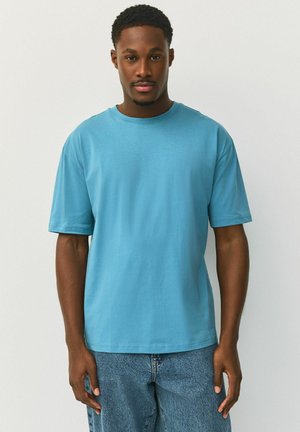 Egy férfi egyszínű kék rövid ujjú pólóban és kék farmernadrágban, egy egyszínű világos háttér előtt áll.