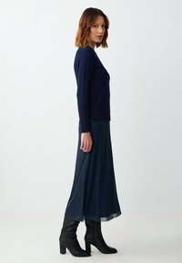 Marineblauwe longsleeve trui gecombineerd met een donkerblauwe gestreepte maxirok en zwarte kniehoge haklaarzen, die een getailleerde silhouet laat zien.