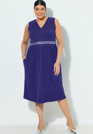 Mujer sonriente, vistiendo un vestido morado sin mangas hasta la rodilla con cintura adornada, manos en los bolsillos, tacones plateados y pendientes de aro, de pie contra una pared blanca.