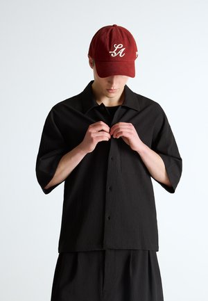 Young person wearing a maroon LA cap buttoning a loose black short-sleeve shirt with matching black pants against a plain background.