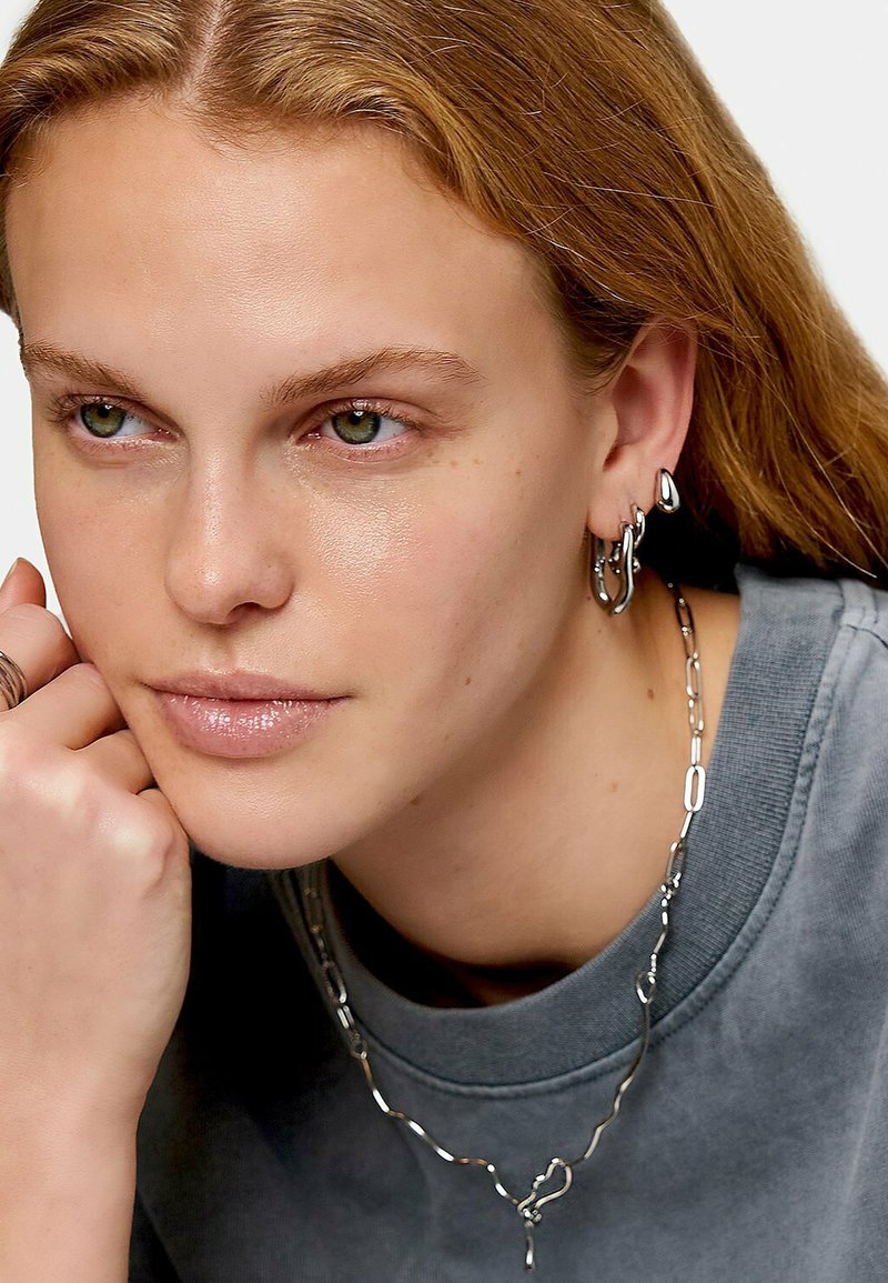Zilveren ketting met onregelmatige schakels, gelaagd met kleinere oorringen. Model poseert met zijn hand op de kin, terwijl ze een grijze shirt draagt.