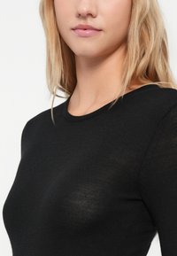 Ξανθιά γυναίκα με μερικώς ορατό πρόσωπο, φορώντας ένα εφαρμοστό μαύρο πλεκτό μπλουζάκι με μακριά μανίκια, με φόντο ένα απλό λευκό φόντο.