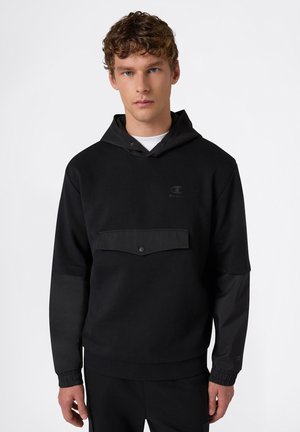 Giovane uomo con capelli ricci che indossa una felpa nera con tasca a patta frontale, in piedi davanti a uno sfondo bianco semplice.