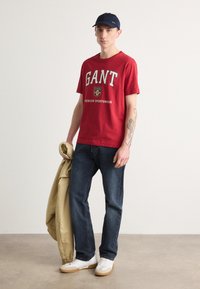 Tricou roșu din bumbac cu logo-ul "GANT", blugi din denim închis, jachetă bej ținută în mâna stângă, adidași albi, pălărie albastră, stând pe un fundal neutru.