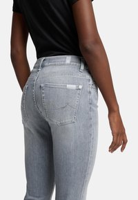Světlé šedé džíny se slim fit střihem, zdobené dekorativním prošíváním na zadních kapsách a zapínáním na knoflík. Bavlněný materiál s hladkou texturou.