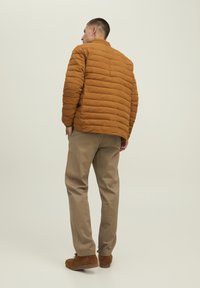 Giacca a vento marrone chiaro con texture a battuta orizzontale, abbinata a pantaloni beige e scarpe in suede marroni. Nessun logo o decorazione visibile.