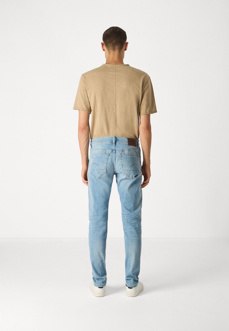 Homme debout de dos, portant un t-shirt beige, un jean bleu clair et des baskets blanches sur un fond blanc uni.