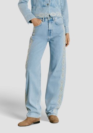 Jeans de mezclilla azul claro con patrones bordados beige a lo largo de las costuras exteriores, combinados con chaqueta de mezclilla a juego y botines de ante marrón.