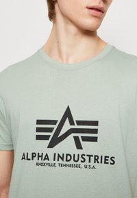 Žalia medvilninė T-maikė su juodu grafiniu logotipu su sparnais ir užrašu "ALPHA INDUSTRIES KNOXVILLE, TENNESSEE, U.S.A."