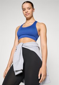 Top desportivo azul com uma textura suave, combinado com leggings pretas. Um moletom cinza claro está amarrado à cintura. Design minimalista.