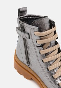 Botín de tobillo sintético gris con un acabado texturizado, cordones beige, cremallera lateral y una suela dura marrón.