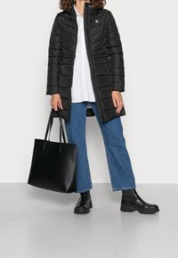 Svart puffjacka med horisontella sömmar, kombinerad med en vit skjorta, blå utsvängda jeans och svarta läderstövlar vid ankeln. Svart tygväska.