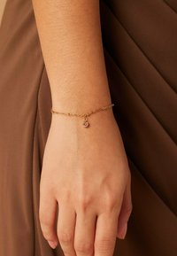 Gouden kettingarmband met een kleine roze edelsteen charm, rustend op een pols met een zachte huid. Het ontwerp is delicaat en minimalistisch.